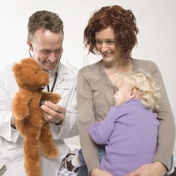 Royalty Free Photo of a Doctor Holding a Stethoscope to a Teddy Bear While a Mother and Daughter Watch