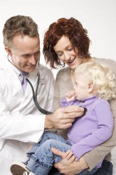 Middle-aged adult Caucasian male doctor holding stethoscope to female toddler's chest with mother watching.