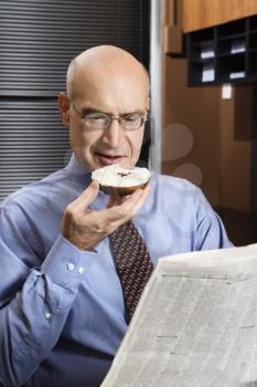 Royalty Free Photo of a Businessman Sitting in an Office Reading a Newspaper While Eating a Bagel