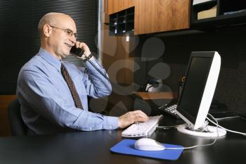 Royalty Free Photo of a Businessman Sitting at Desk in an Office Talking on the Telephone and Typing on a Computer