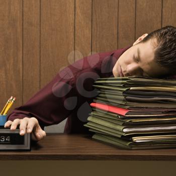 Royalty Free Photo of a Businessman Sitting at a Desk With Head Down Sleeping on a Tall Stack of Folders
