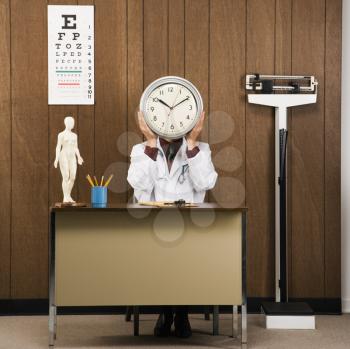 Royalty Free Photo of a Male Doctor Sitting at a Desk Holding a Clock Over His Face
