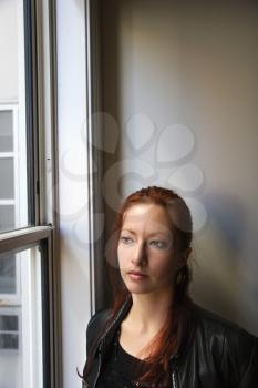 Pretty redhead young woman standing indoors by window looking out.