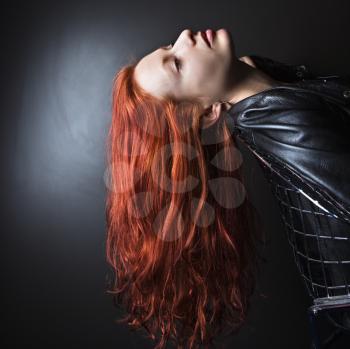 Pretty redhead young woman wearing leather jacket leaning back in chair with long hair hanging down.