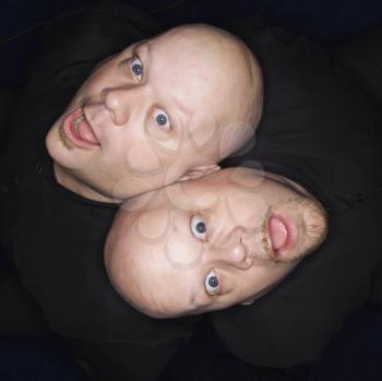 Aeriel view of Caucasian bald identical twin men sitting back to back making facial expressions.