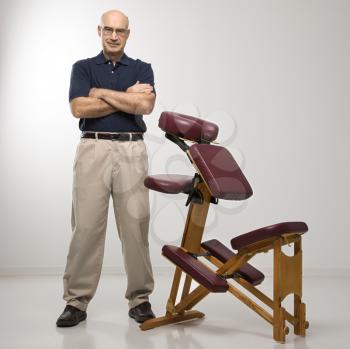 Caucasian middle-aged male massage therapist standing with arms crossed beside massage chair.