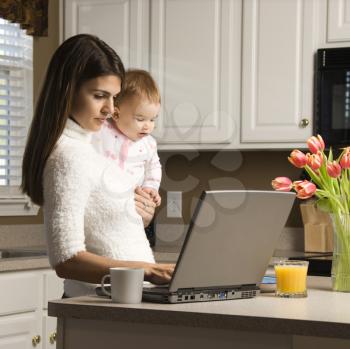 Royalty Free Photo of a Mother Holding Her Baby and Typing on a Laptop in the Kitchen