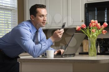 Royalty Free Photo of a Man in a Suit Using a Laptop Computer and Cellphone in a Kitchen