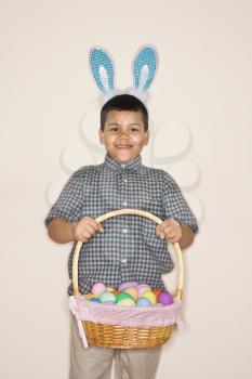 Hispanic boy wearing bunny ears holding Easter basket smiling and looking at viewer.