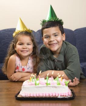 Royalty Free Photo of a Girl and Boy Wearing Party Hats Sitting in Front of a Birthday Cake Smiling