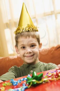 Caucasian boy wearing party hat holding gift  and smiling at viewer.