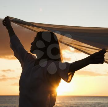 Royalty Free Photo of a Woman Holding Up Fabric in the Breeze Silhouetted by Sunset Beside Ocean