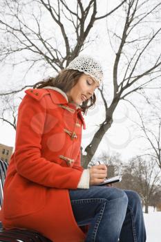 Low angle view of Caucasian young adult female in winter clothing sitting on park bench using PDA .
