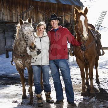Royalty Free Photo of a Couple Holding Horses in Winter with a Stable in the Background