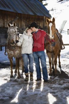 Royalty Free Photo of a Couple Kissing Holding Horses in Winter with a Stable in the Background