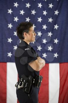 Side view of mid adult Caucasian policewoman standing with arms crossed and American flag as backdrop.