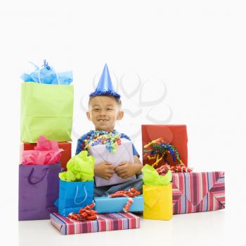 Asian boy sitting smiling wearing party hat with wrapped presents.