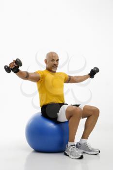 Mid adult multiethnic man balancing on blue exercise ball with outstretched arms holding dumbbells and looking at viewer.