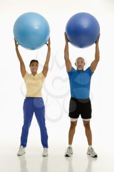 Smiling mid adult multiethnic man and woman holding blue exercise balls over their heads looking at viewer.