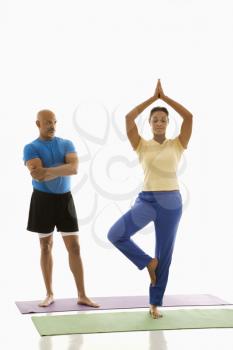 Mid adult multiethnic woman balancing in tree yoga pose while mid adult multiethnic man stands and watches.
