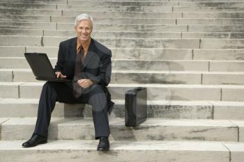 Caucasian middle aged businessman sitting on steps outdoors with laptop and briefcase smiling at viewer.