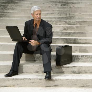 Royalty Free Photo of a Middle-Aged Businessman Sitting on Steps Outdoors With a Laptop and Briefcase