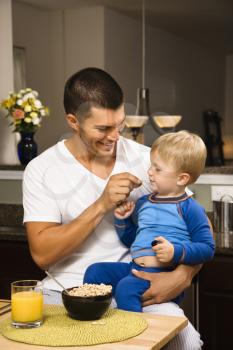 Caucasian man feeding toddler son on lap in kitchen. 