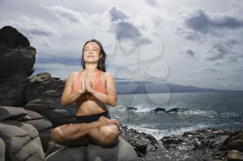Royalty Free Photo of an Asian Woman Sitting on a Rock by the Ocean in Lotus Pose with Eyes Closed in Maui, Hawaii