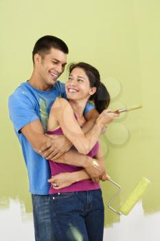 Royalty Free Photo of a Couple Smiling and Embracing in Front of a Partially Painted Wall Holding a Paintbrush and Roller