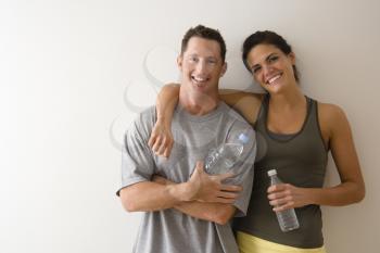 Man and woman at gym in fitness attire holding water bottles standing against wall smiling.