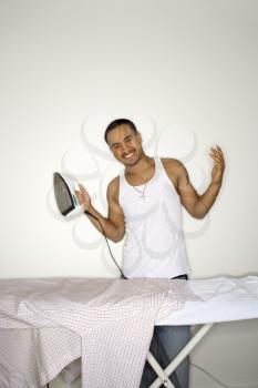 Happy young man standing behind an ironing board. He is shrugging and holding an iron. Vertical shot.