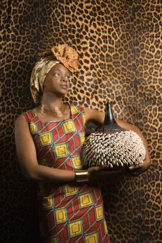 Portrait of an African American woman wearing traditional African clothing and holding a shekere in front of a patterned wall. Vertical format.