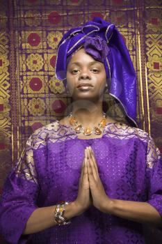 Portrait of an African American woman wearing traditional African clothing in front of a patterned wall and holding her hands in a prayer position. Vertical format.