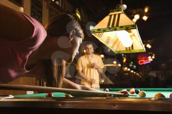 Young woman playing billiards with a young man in the background.  Horizontal shot.