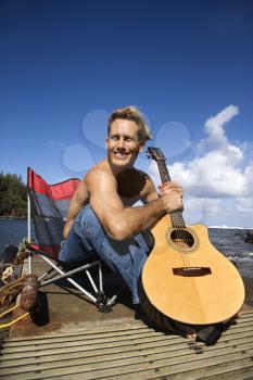Young man sitting on pier shirtless holding a guitar and smiling. Vertical shot.