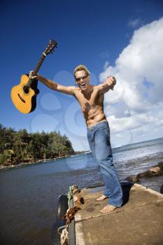 Shirtless young man wearing sunglasses, standing on a pier and holding a guitar up in the air with an enthusiastic expression. Vertical shot.