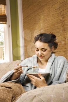 Young woman sitting on a couch in a bathrobe eating soup. Vertical shot.