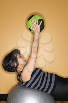 Asian woman performs a conditioning exercise with a medicine ball while leaning back on a balance ball at the gym. Vertical shot.