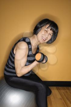 Asian woman sits on a balance ball while lifting a hand weight. She is smiling towards the camera. Vertical shot.