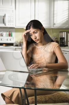A young woman sits at the kitchen table using a laptop. She has a worried expression on her face. Vertical shot.