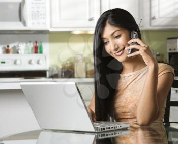 A young woman chats on her mobile phone while sitting at the kitchen table in front of a laptop.  Horizontal shot.