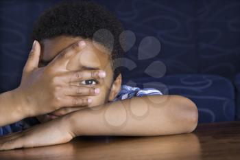 Young African American boy peeks through his fingers as he sits at a coffee table. Horizontal shot.