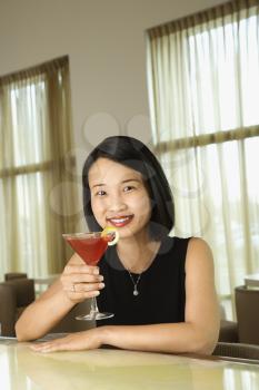 Attractive young Asian woman smiling and holding up a cocktail at a bar. Vertical shot.