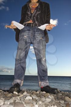 A young man poses at the beach. He is viewable from the neck down, holding a cigarette, and is showing his empty jeans pockets. Vertical shot.