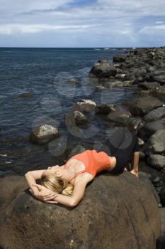Attractive young woman lies on top of a large rock on a rocky coast. Vertical shot.