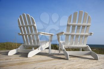 Rear view of two white Adirondack style chairs sitting on a wooden deck, facing the beach. Vertical shot.