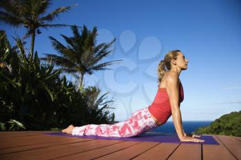 Attractive young woman in red practices yoga on a deck with the ocean in the background. Vertical shot.