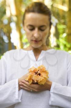 Attractive woman in white holds a hibiscus flower in her hands as she meditates. Vertical shot.