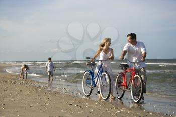 Husband and wife walk their bikes down the beach with children in the background. Horizontal shot.