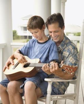 Son sits on his father's lap while playing guitar. Vertical shot.